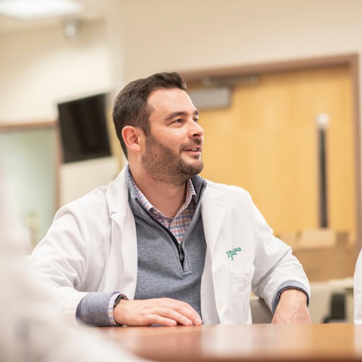 A nurse in a white lab coat smiles as he looks forward. He is seated around a table with other medical professionals in lab coats visible in the foreground.