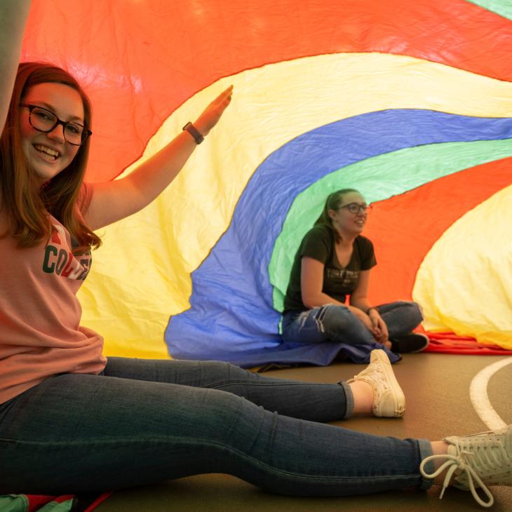 A group of students sit on the floor holding up a large circular, multi-colored sheet over them.
