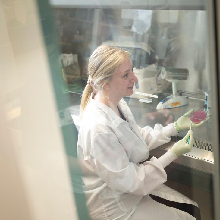 A student holds a petri dish with a sample in one hand and an inoculating loop in the other.