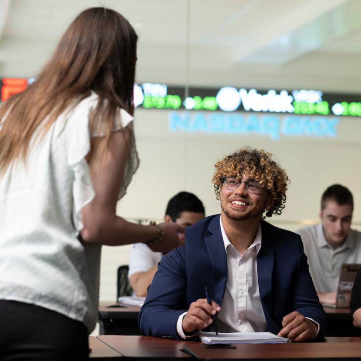 A professor speaks to smiling students in a business classroom. A NASDAQ ticker is visible in the background.