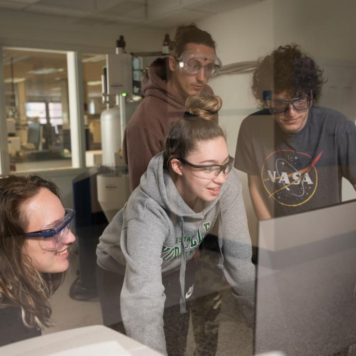 A group of 4 students, all wearing safety goggles, gathers around a large monitor in a room full of chemistry lab equipment and large machinery.