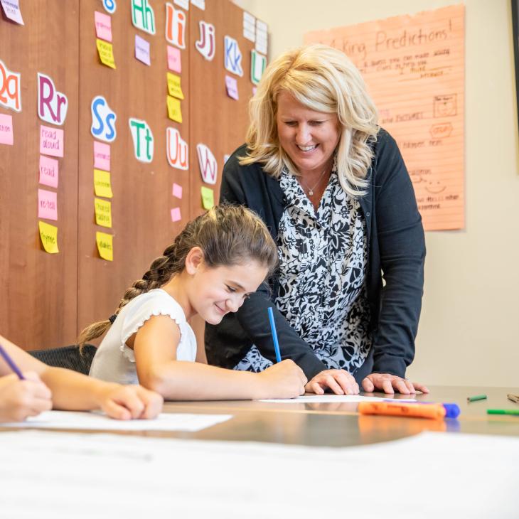 A teacher leans forward on a table as a young student works on a project with a blue colored pencil.