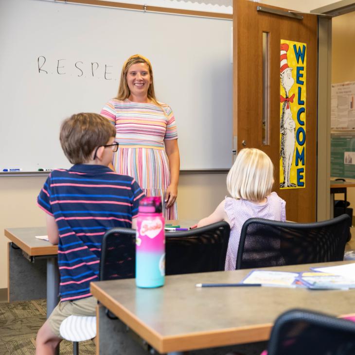 A teacher stands in front of the whiteboard at the front of an elementary classroom. Two students are visible, seated at desks.