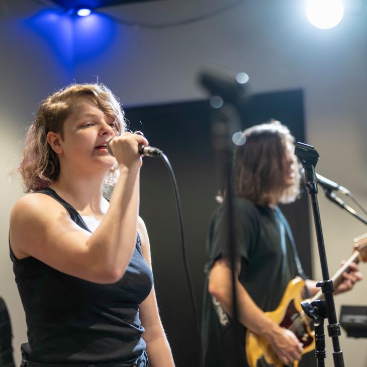 Two students sing into standing microphones. One of the students plays an electric guitar.