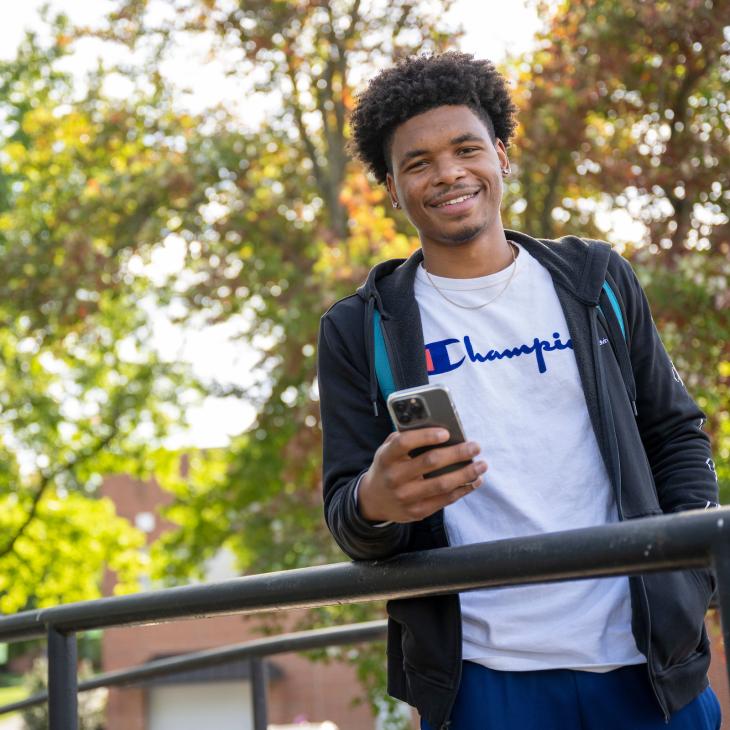 Student on a bridge smiling and holding a phone, wearing a black coat over top of a white shirt.