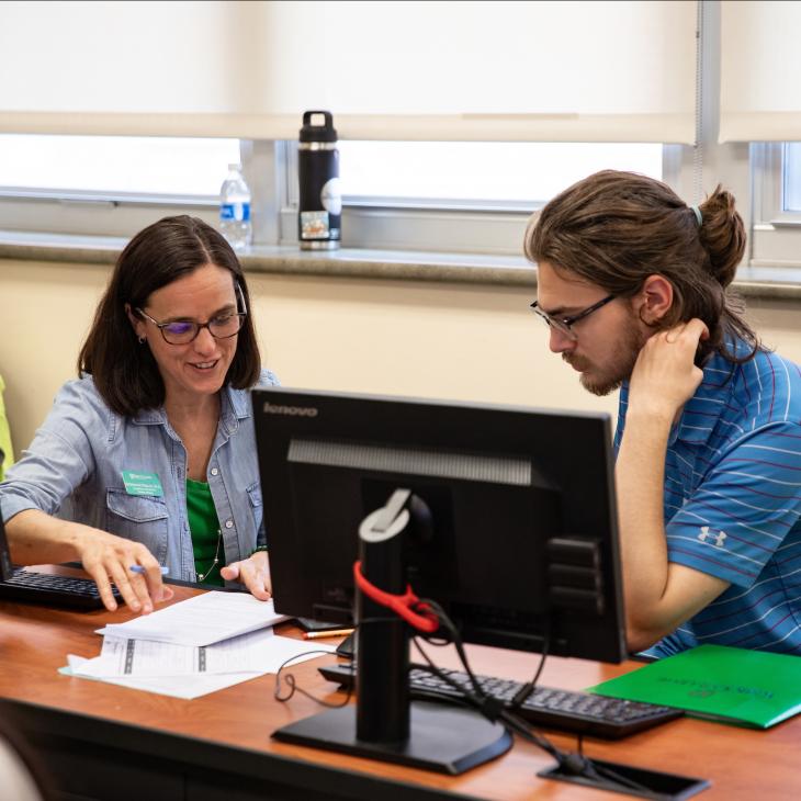 A staff member kneels beside a student at a computer lab desk, helping with scheduling. 