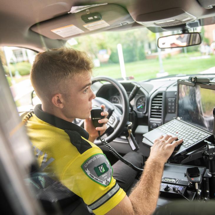 A student Campus Safety officer answers a radio call in a patrol car.