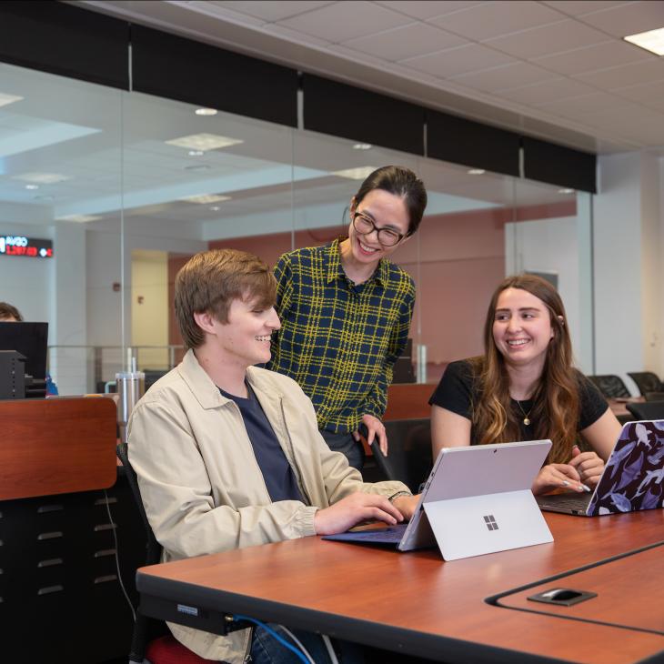 Two students look on from a classroom desk as a professor stops to chat with them. A tablet rests on the table in front of them.