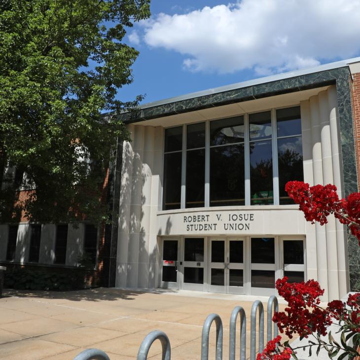 A view of the Iosue Student Union building on a sunny day. The entrance is framed by large green trees at left and red flowers at right.