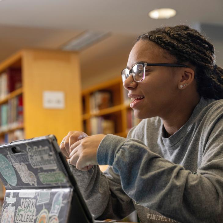 A student sits in the library, surrounded by bookshelves as she works on an iPad.