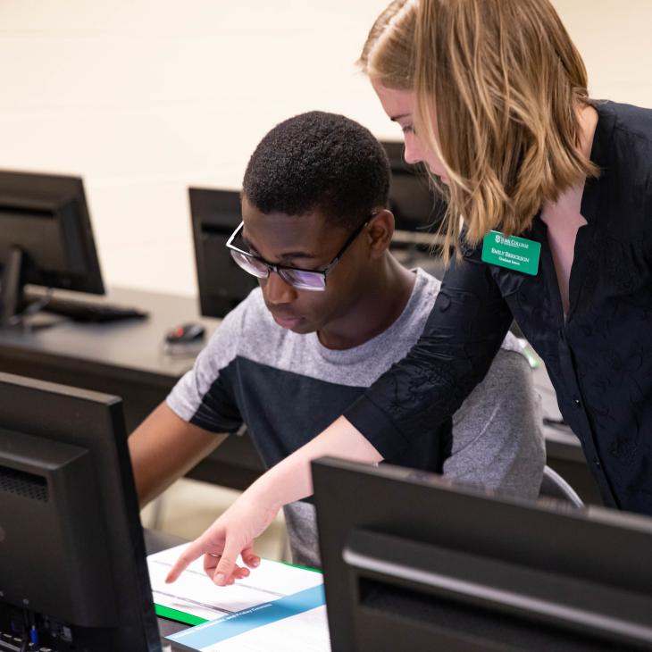 A student works on a computer in the computer lab as an advisor points at the screen to provide instruction.