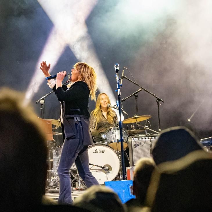 A side view of a lead singer for a band at the White Rose Music Fest. The drummer is seated and playing behind the lead singer, and crossed spotlights are shining up behind the band.