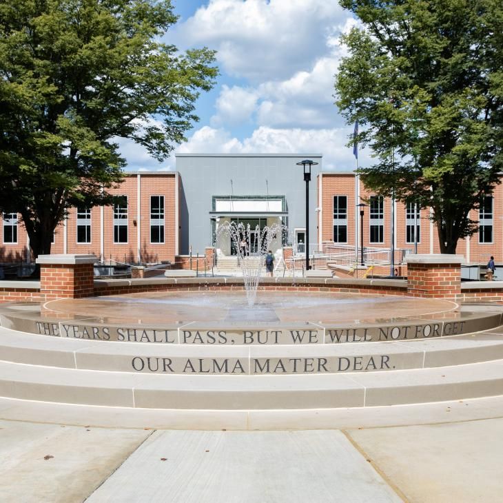 A view of the campus fountain flowing in front of the Schmidt Library. The fountain flows from the top of a three flat concrete tiers and is framed by a concrete topped, staggered brick wall.