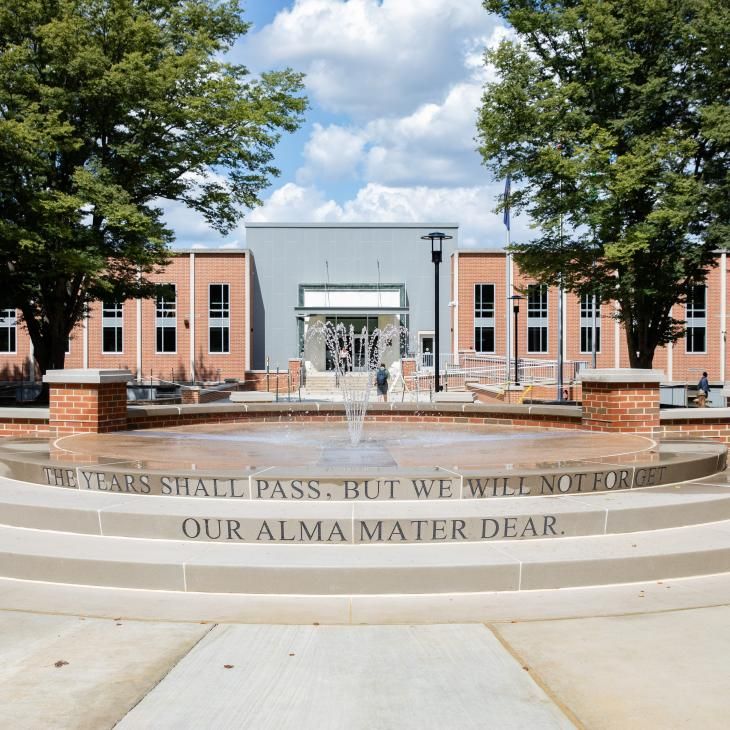 A view of the campus fountain flowing in front of the Schmidt Library. The fountain flows from the top of a three flat concrete tiers and is framed by a concrete topped, staggered brick wall.