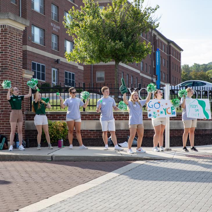 Group of students outside of a residence hall cheering and celebrating new students moving in.