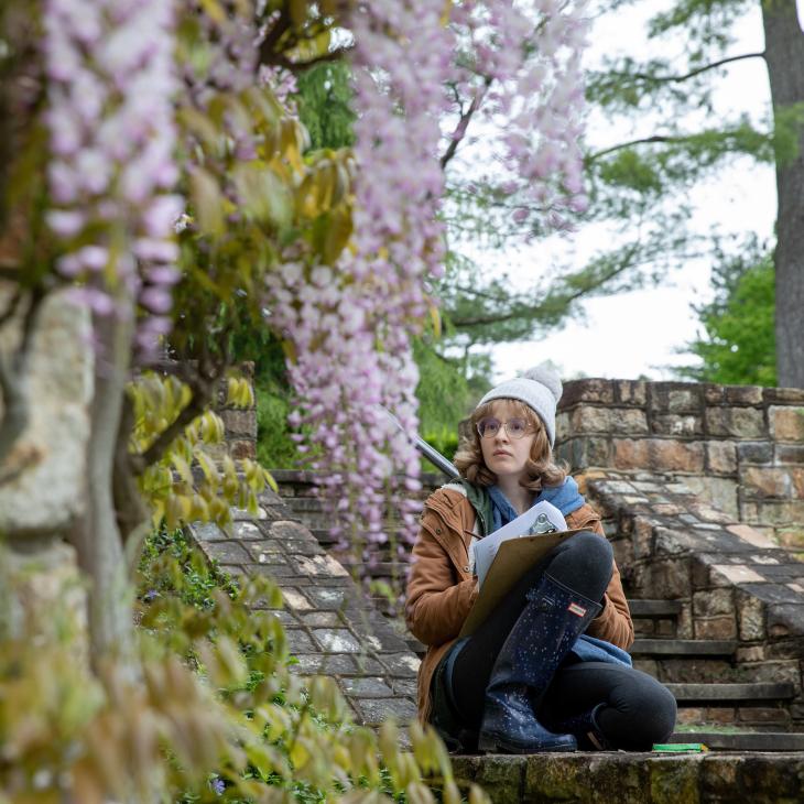 A student wears a winter coat and hat while observing ornamental plants and taking notes on a clipboard.