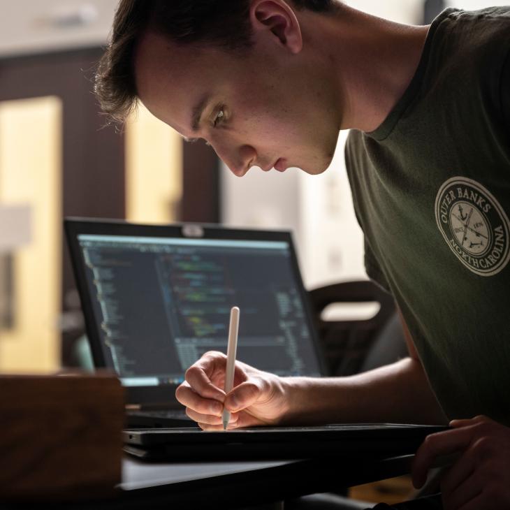 A student writes on a tablet with a digital pencil in a computer lab. An open laptop is visible on the desk beside him.
