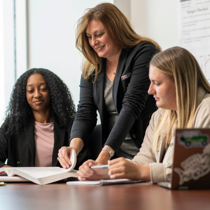 Professor Renee Tacka looks through a textbook with two students in a classroom.