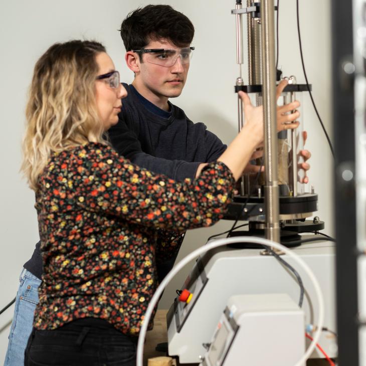 A professor shows a student how to use a piece of machinery in the engineering lab. Both wear safety goggles.