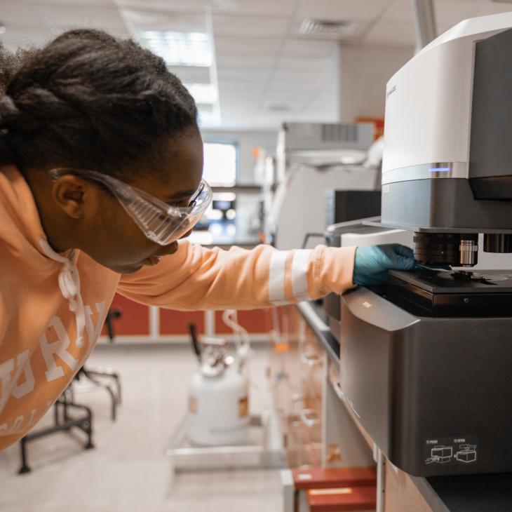 A student wearing gloves and safety goggles works with a machine in the chemistry lab.