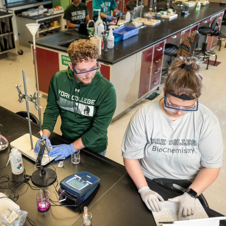 Two students work in a chemistry lab, surrounded by equipment.