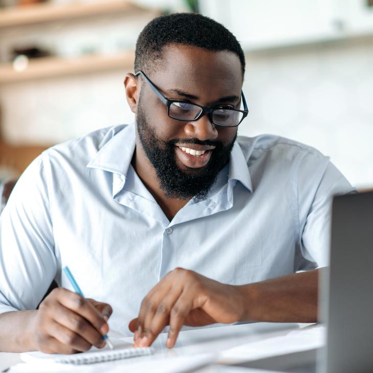 A man in glasses with a pen in his hand writing while looking at a laptop screen.