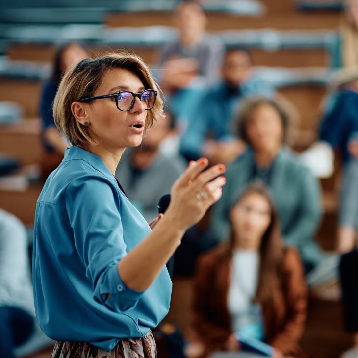 A woman speaking in front of a group in a large room.