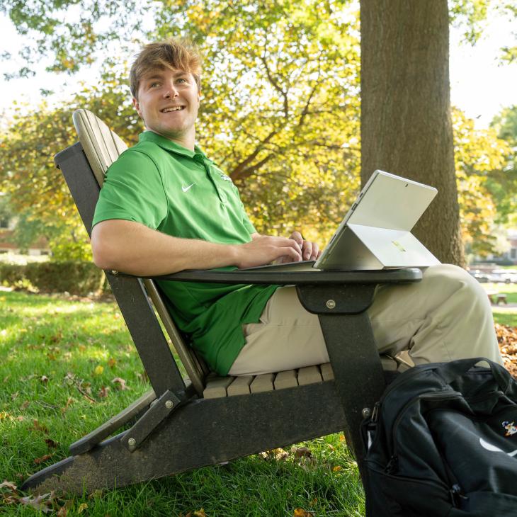 Student in green polo sitting outside in a chair looking at an iPad