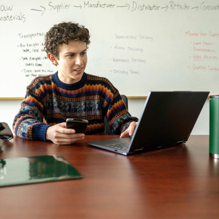 Student holding calculator and looking at laptop at a desk