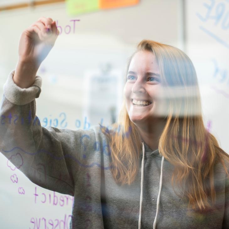 Student writing on clear board while smiling