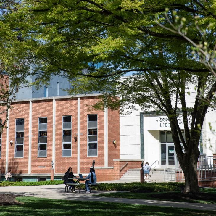 Two students sit in the shade at a picnic table in front of the York College library.