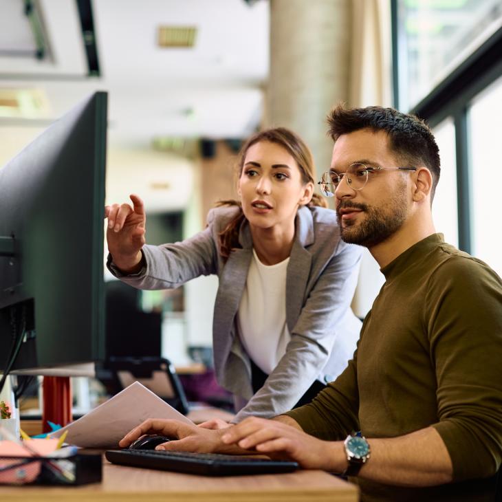 Two professionals look at a large desktop computer monitor in an office setting.