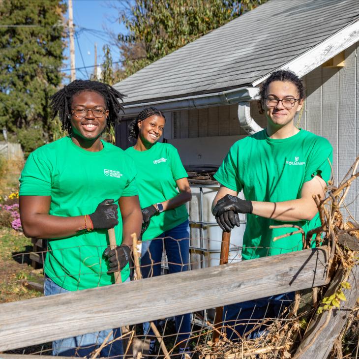 Three students in green t-shirts smile at the camera while working in a garden