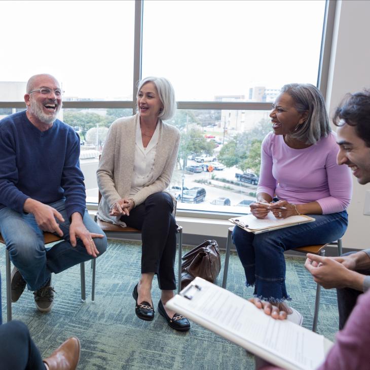 A group of diverse older adults laughing together in a room with large glass windows behind them.