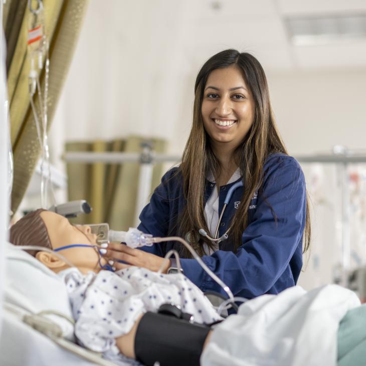 Nursing major evaluates a patient in a simulation lab, smiling to camera while holding an intubation tube near a patient bed.
