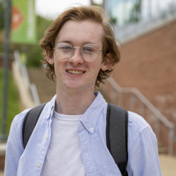 College student with glasses smiles and a backpack smiles at the camera with the exterior of a building and a set of stairs visible in the background.