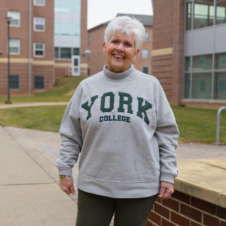 An older woman in a York College sweatshirt stands outside of tall brick buildings