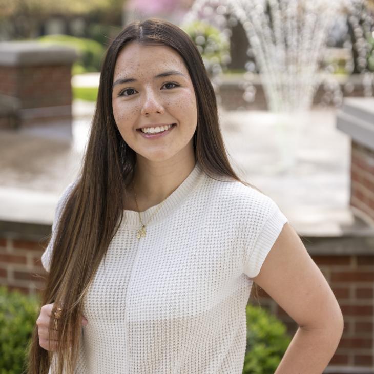 Portrait of Supply Chain major Megan Holtzinger '26, smiling at the camera on a sunny day with a fountain and trees visible in the background.