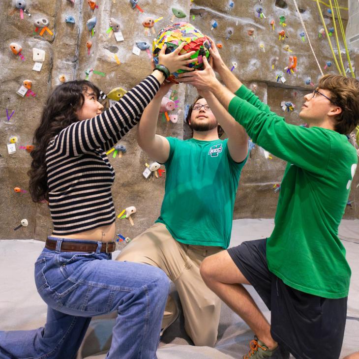 Three people kneeling in a rock climbing gym, holding a colorful ball of tape overhead.