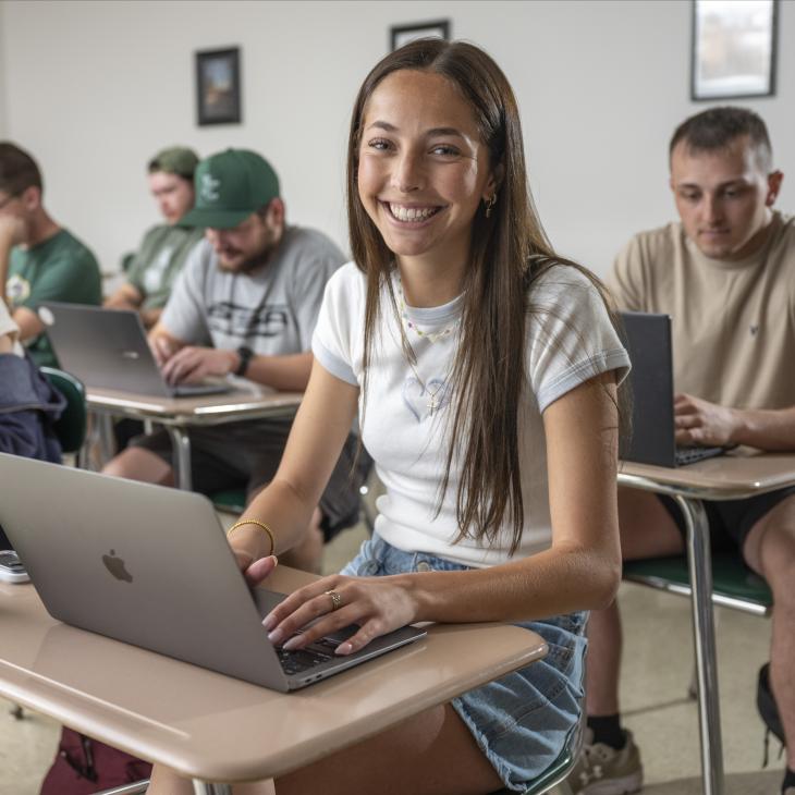 Students sitting in a classroom with their laptops open. A single student smiles at the camera.