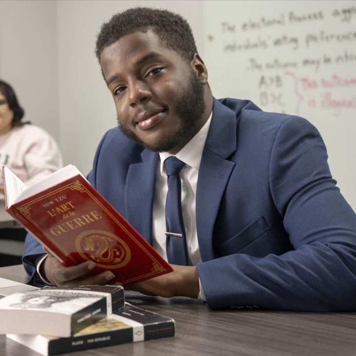 A student in a suit reads a book with a red cover in front of an open laptop.