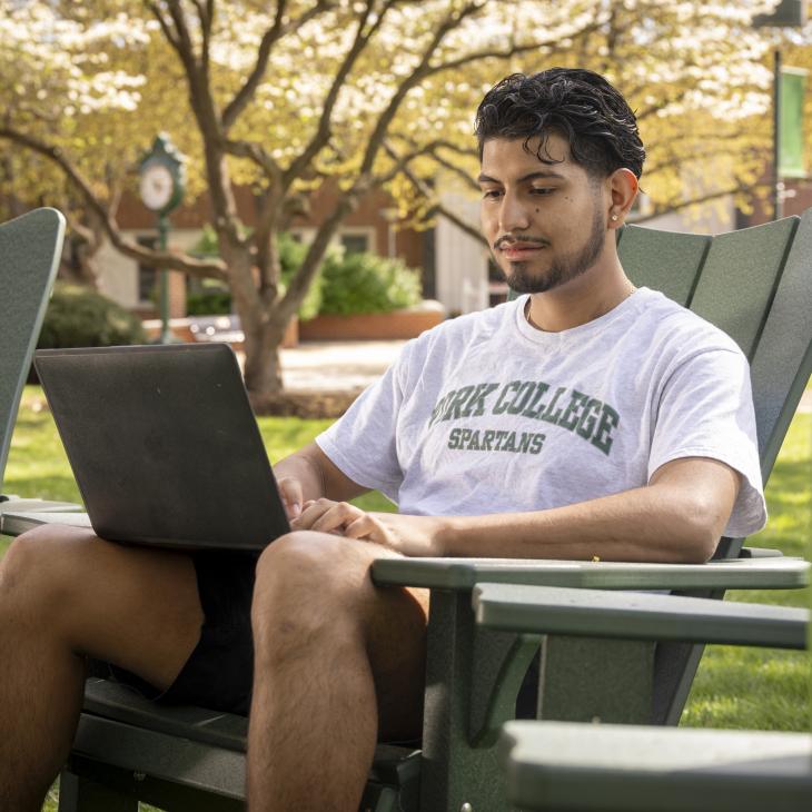 A student wearing a York College t-shirt works on a lap top while sitting outside on an Adirondack chair.