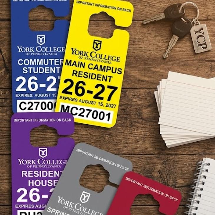 An overhead view of five colorful York College of Pennsylvania hanging parking permits for 2026–2027 arranged on a wooden desk alongside car keys with a YCP keychain, index cards, a spiral notebook, and eyeglasses.