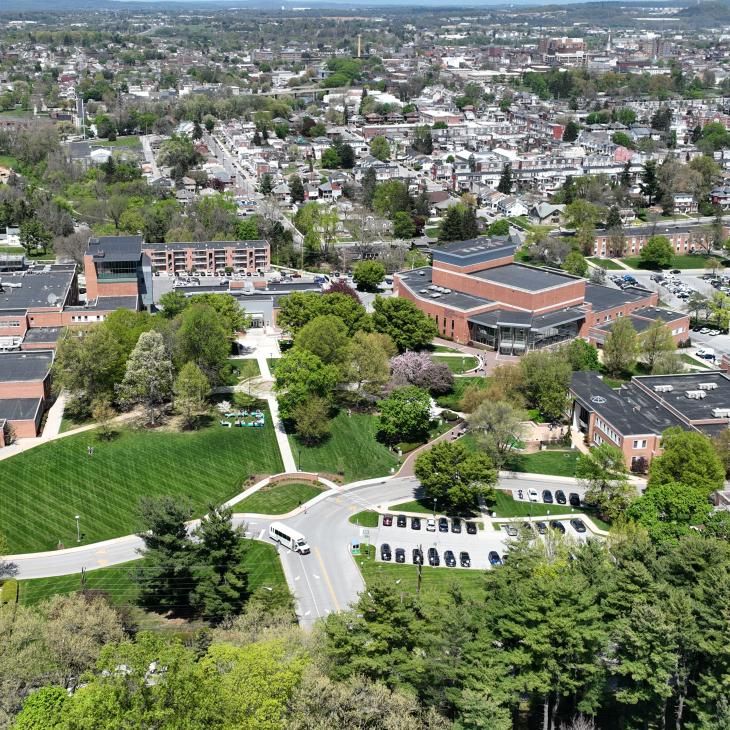Aerial view of a college campus with red-brick buildings, green lawns, and surrounding residential neighborhoods under a clear blue sky.