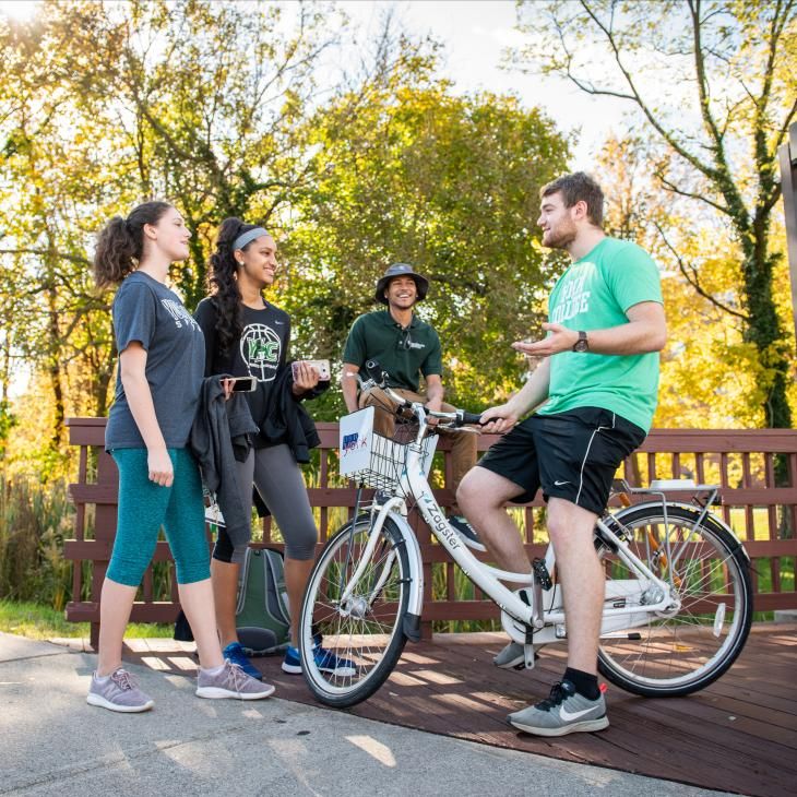 Students talking with a cyclist on a campus walkway during a sunny fall day.
